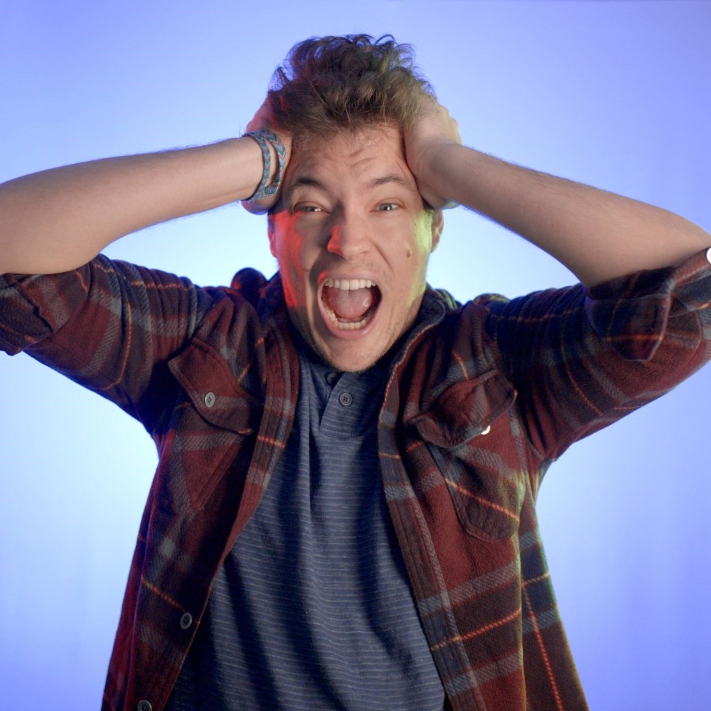 Photo of Austin on a light blue background, screaming and holding his arms to his head in a humorous attempt at horror.
